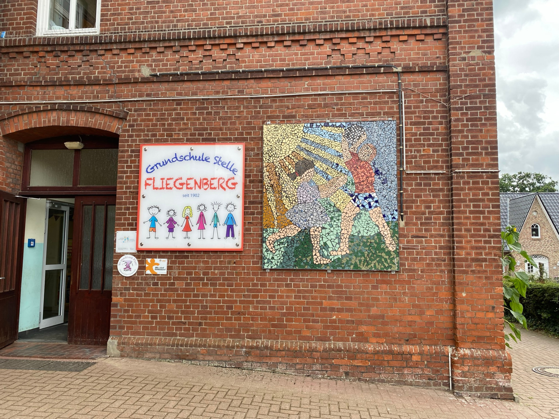 Mosaikbild an der Klinkerfassade neben dem Schulschild „Grundschule Stelle Fliegenberg, seit 1902“